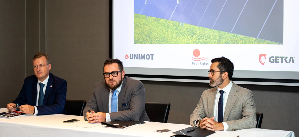 A white-collar worker in formal wear holding a microphone at an event featuring clothing, outerwear, and electronic devices. UNIMOT, GETKA, and First Solar logos featured prominently.